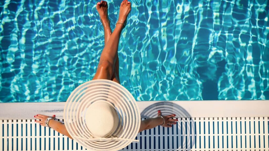Mulher com chapéu de praia sentada na beira da piscina.